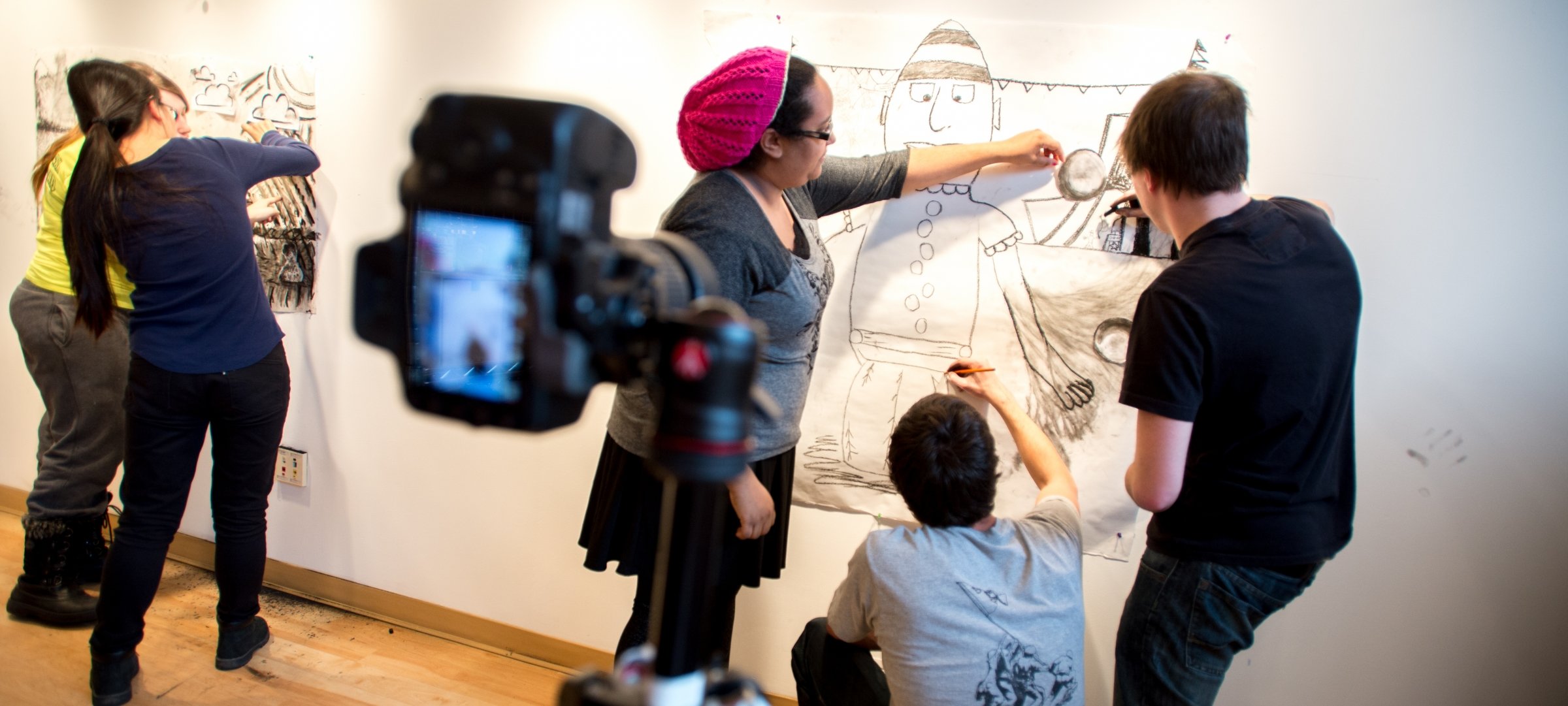Faculty works with students on a large drawing on a wall with a camera looking on.