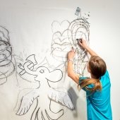 Student taping a large drawing to the studio wall as another photographs