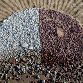 Pebbles on a sandy beach in a semi-circle, lighter pebbles on the left, darker on the right, with a clear vertical division line. The pebbles are concentrated at the top of the semi-circle and trickle down to less concentrated stones.