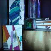 Seven boxes with abstract paintings on their faces. The first box has two pyramids with various patterns. The second box, stacked on top of the first box, contains the outline of a person, with a striped blue and green pattern. The third box contains golden rays of light with white highlights and particles. The fouth box, stacked on top of the third box, has black shapes with green outlines. The fifth box has