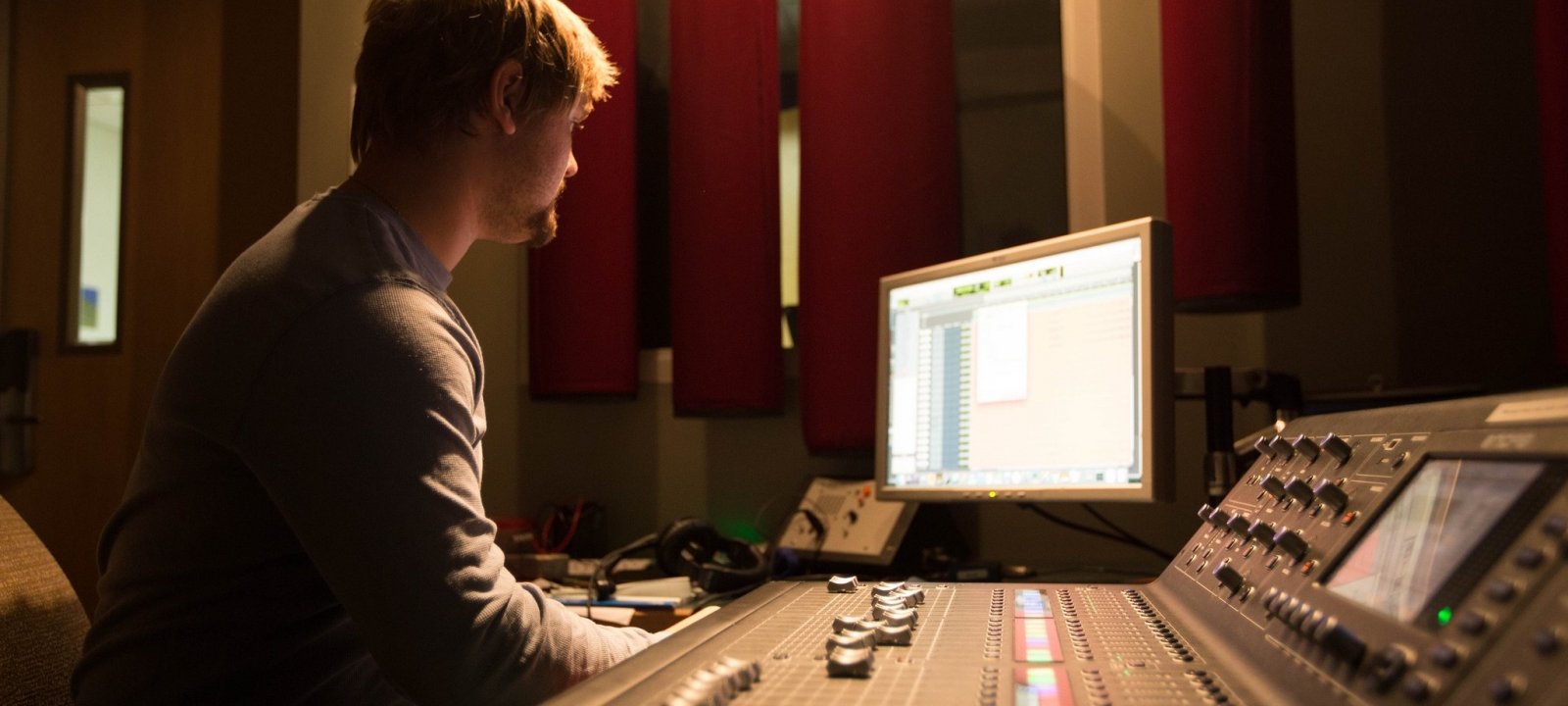 Student sitting inside the sound booth at a computer and console making adjustments during a performance.