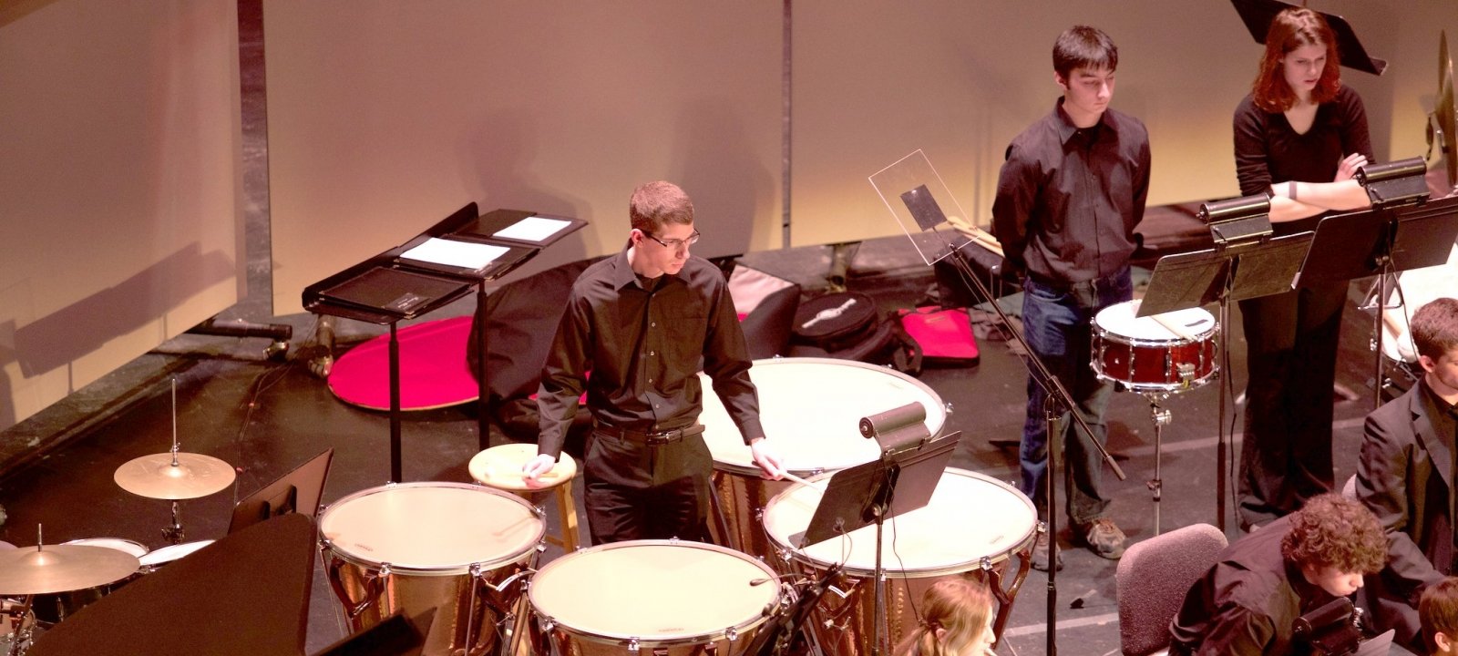 Student in a concert band standing in the middle playing large drums while looking a music on a music stand during a performance.