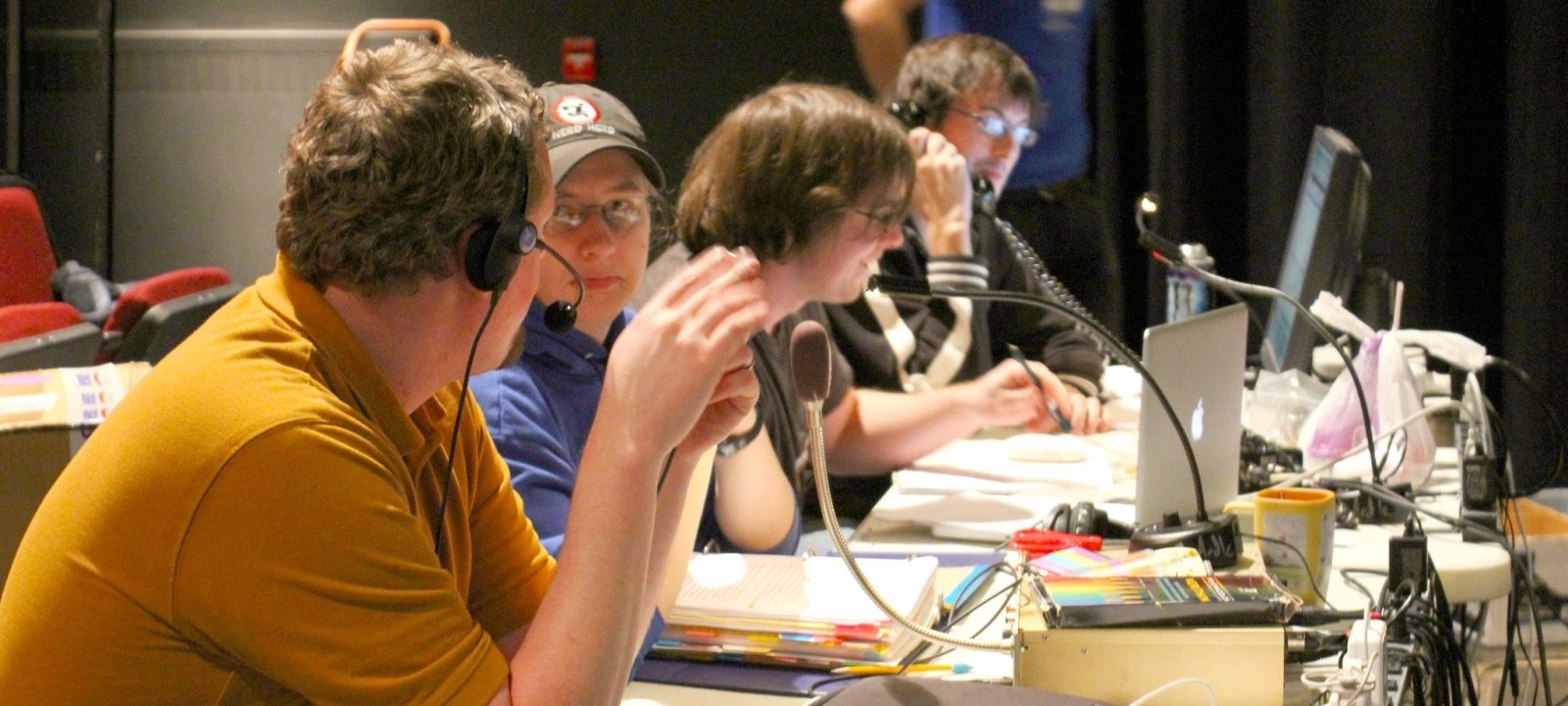 Students sitting backstage at a table with laptops, they have headsets and microphones on and are discussing with each other.