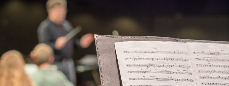 A music stand with sheet music. In the background, Joel Neves leads the Keweenaw Symphony Orchestra in performance. 