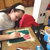 Student sitting, leaning over a painting with a brush.