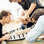 Two students looking on as a third knees down to drill into a wooden block on a circular project.