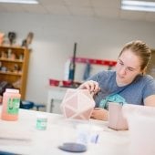 Student sits at a workbench painting a sculpture.