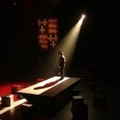 An actor stands alone in a knight's costume with a single light shining on the stage.