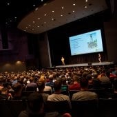 Audience watches two speakers and a presentation in the Performance Hall.