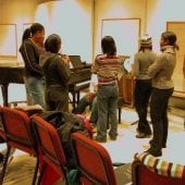 African American visiting harp student group surrounds someone seated at a piano while one writes on a lined dry erase board music notes.