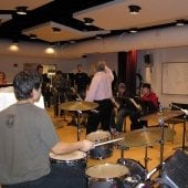 Students seated with instruments behind music stands with music looking to the instructor in the center of the room for guidance.