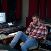 Single student sitting in a chair in front of monitor and sound board with hand under chin and leg propped up.