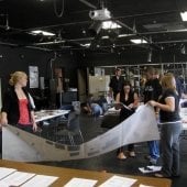 Students holding a large poster while two student confer over a binder inside the Lighting and Sound Lab.