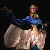 Model posing and extending her arms in a colorful costume with newsprint wings on her arms held with string.