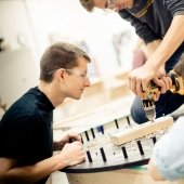 Two students observe as a third drills into a block of wood on a round, pegged project.