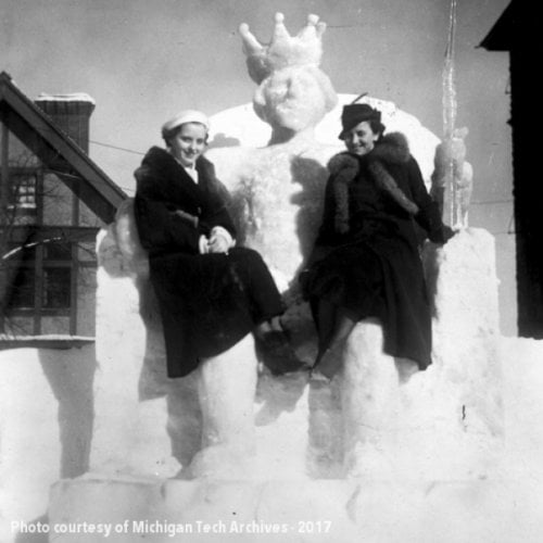 two women sit on the lap of a King Neptune ice and snow sculpture.