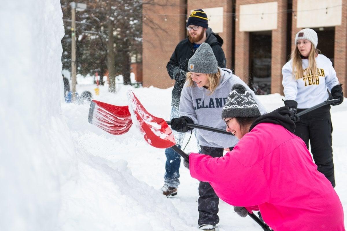 Three young women and a young man pile snow outside. One woman is wearing a Michigan Tech sweatshirt and another is wearing a Leland shirt, with a brick building in the background on a college campus.