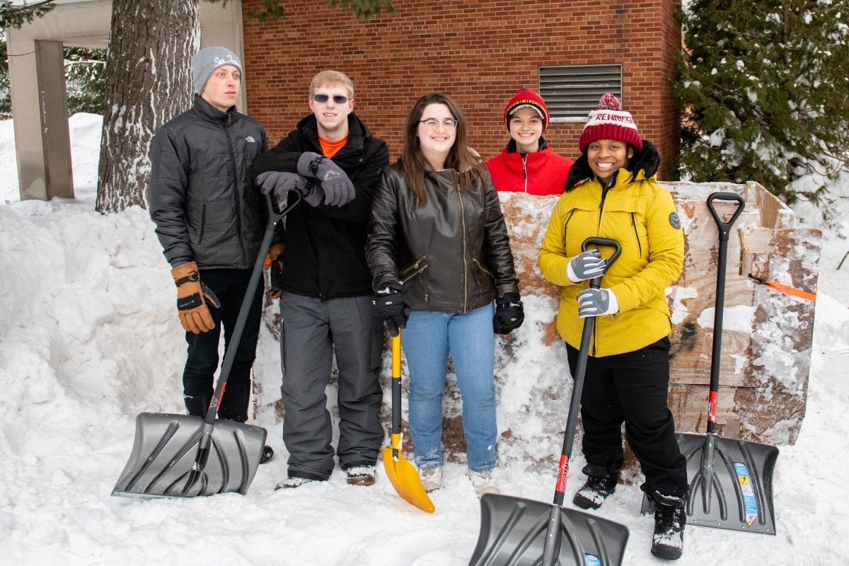 Five students stand with shovels on a college campus with a plywood statue form under a tree in front of a brick building for Winter Carnival.