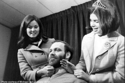 A man with a beard sits between two women in the 1970s in a black-and-white photo inside during a beard judging contest. The woman with the crown and the other woman are checking the man's beard for softness.