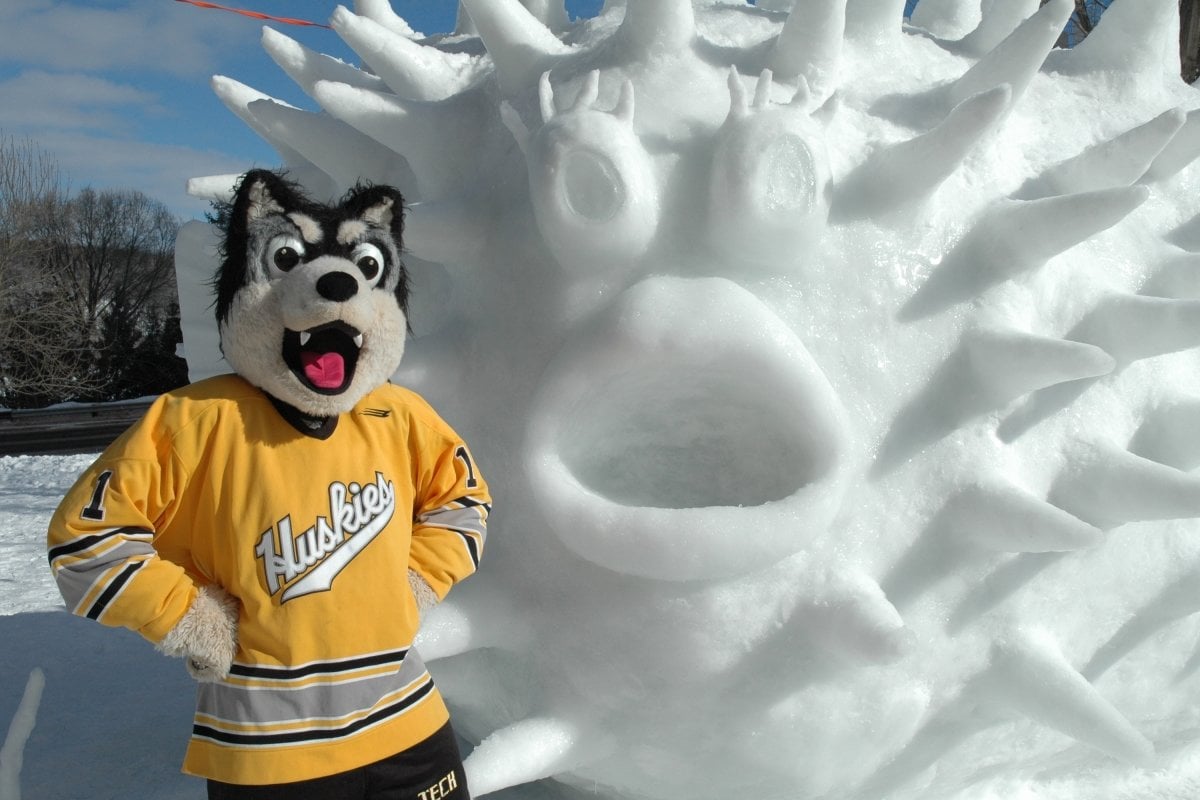 A Husky mascot makes a round O of his mouth copying a fish statue made of ice and snow outside in the winter.