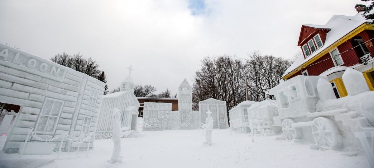 Wide-angle of a snow statue with a saloon and wild west figures with an old yellow and burgandy mansion in the background.