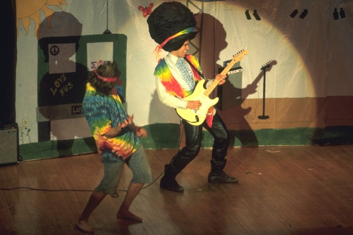Two students on a stage wear tie-dyed accessories. One plays guitar wearing a giant wig; the other plays air guitar.