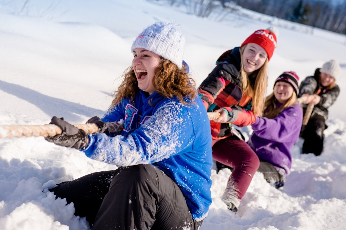 Four young women hold a thick rope on their knees in the snow in a tug-of-war-game outside with a blue sky and forest treeline in the background.